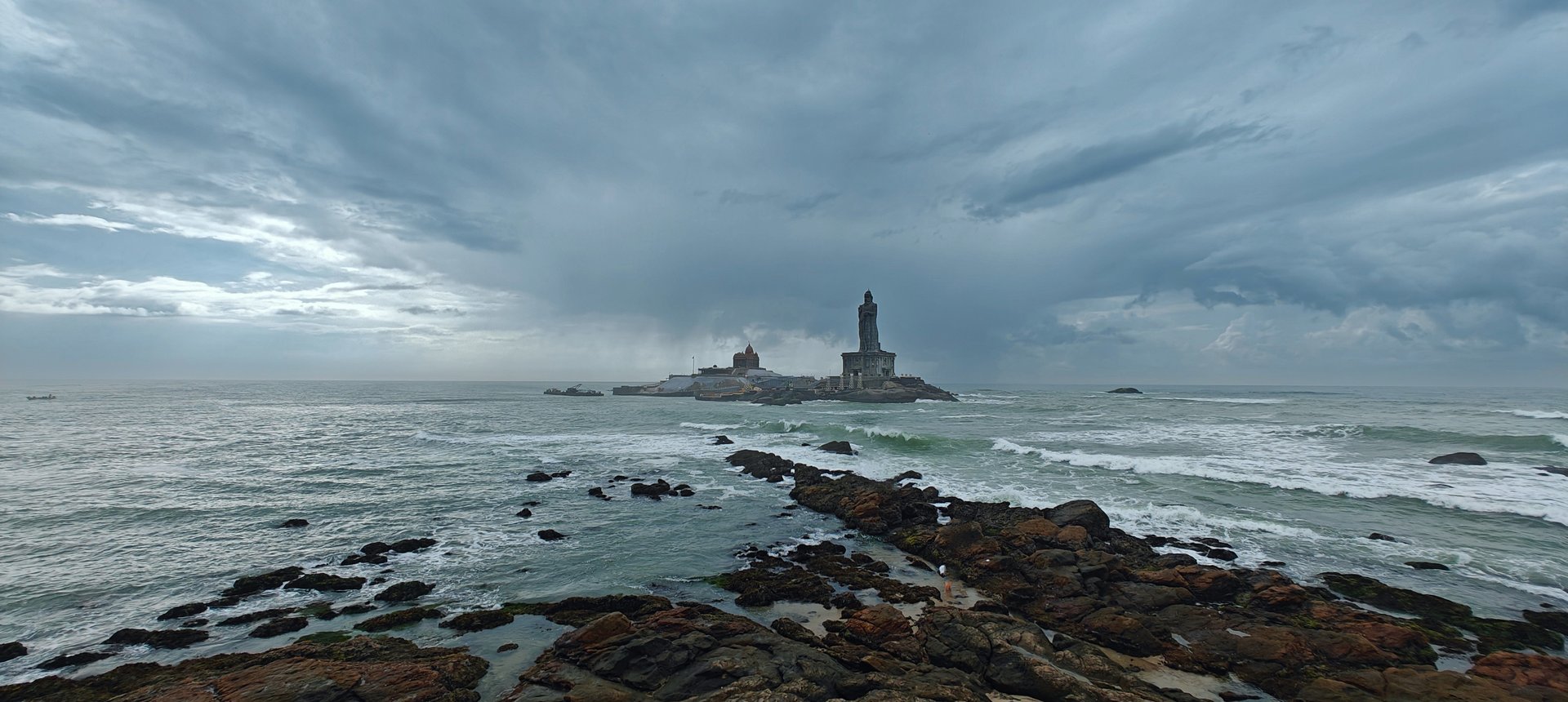 Vivekananda Rock Memorial and Thiruvalluvar Statue seen from the rocky shores of Kanyakumari, with dramatic storm clouds over three oceans — Photo: Sabin, sabinv.com